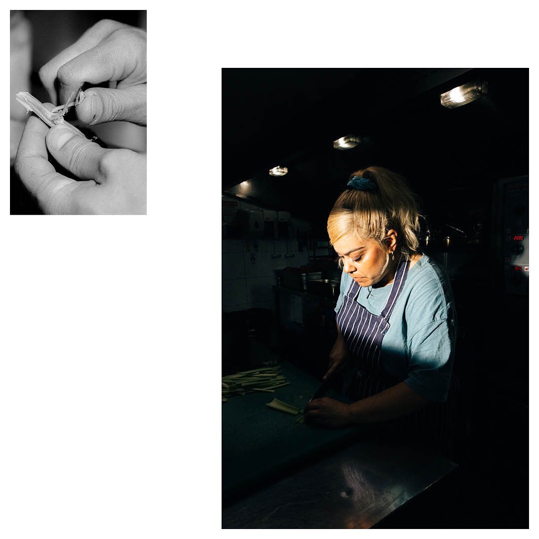 Chef at work in dark kitchen, preparing food. Woman wearing blue shirt and apron, blonde hair in ponytail, lights overhead.