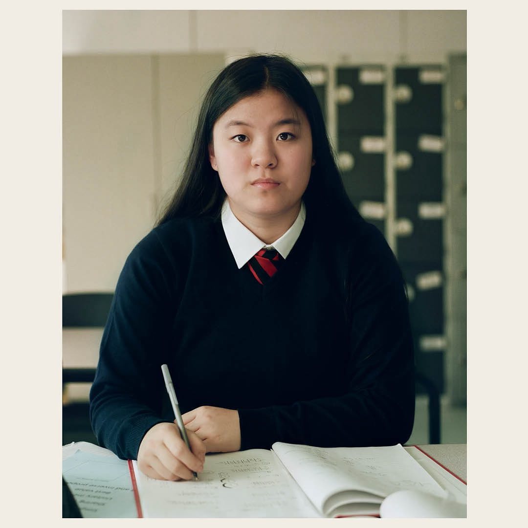 Portrait of an Asian teenage girl in a school uniform sitting at a desk, writing in a notebook, with lockers in the background.