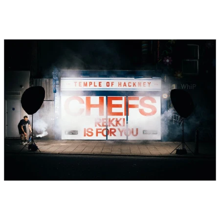 Night shoot with a person holding a smoke emitter in front of building with large text and softbox lighting in an urban environment.