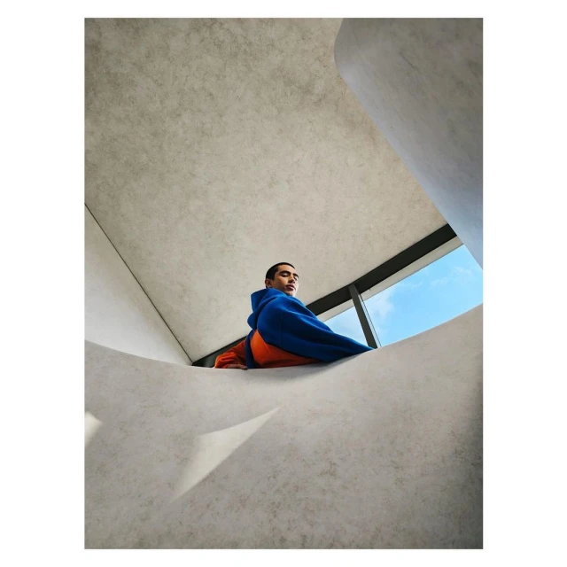 Stylish adult male in blue hoodie and orange shirt poses on modern concrete architecture with blue sky visible through window opening.