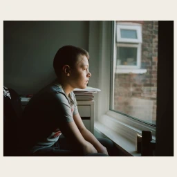Young boy seated indoors looking thoughtfully out a large window by Aria Shahrokhshahi