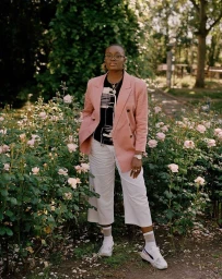 Portrait of a Black woman in a pink blazer standing amidst blooming rosebushes in a park, captured by Aria Shahrokhshahi.
