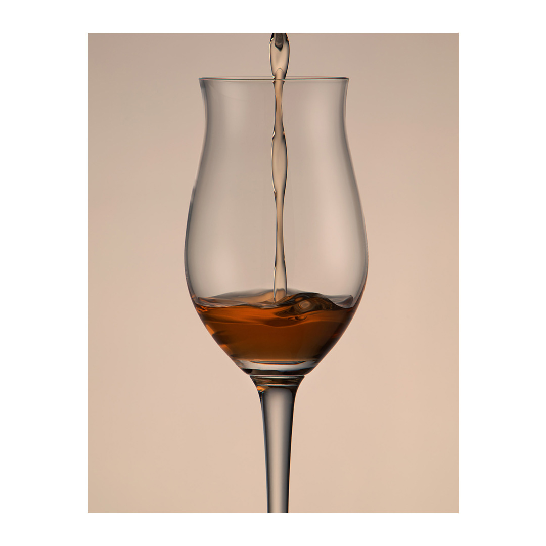 Amber liquid is poured into an elegant, clear stemmed glass, creating a visual display of motion and reflection against a neutral backdrop.