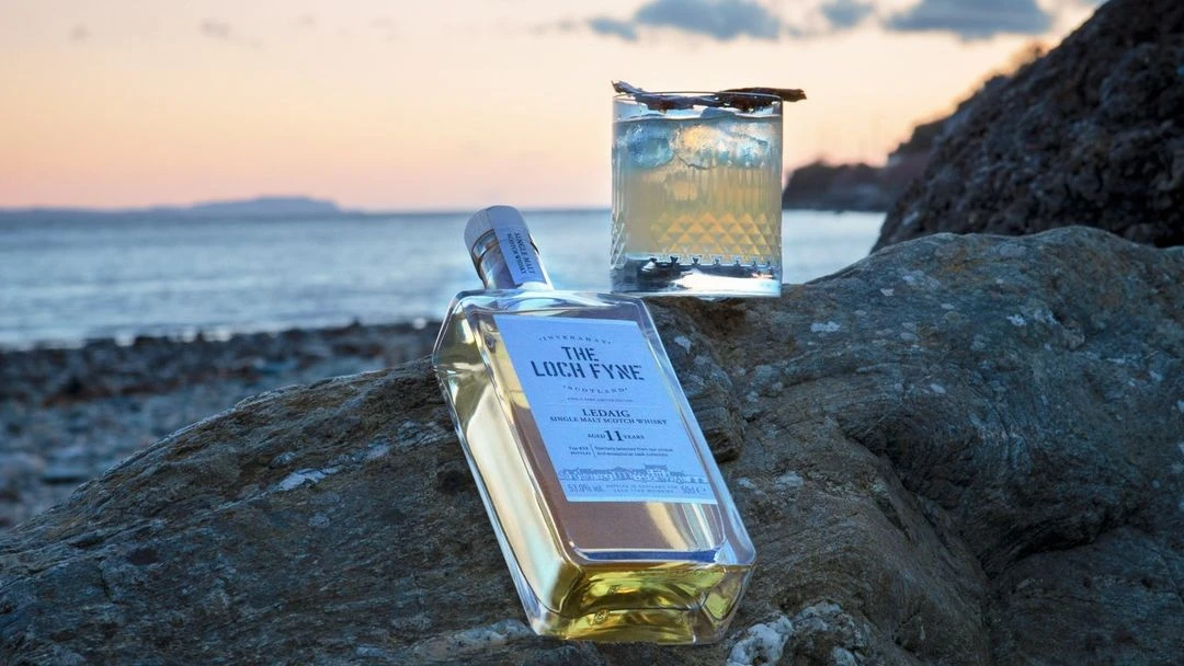Whiskey bottle and cocktail glass arranged on a rock near the ocean during sunset, creating a scenic and inviting beverage still life.