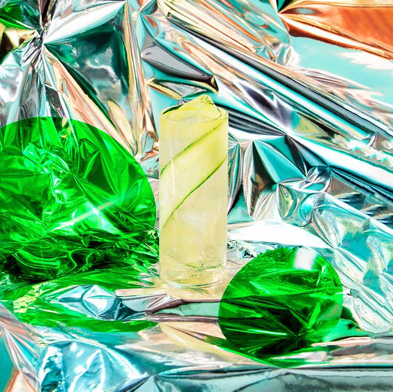 Tall glass of cucumber-infused beverage with ice, set against a reflective silver background with green accents, vibrant and refreshing.