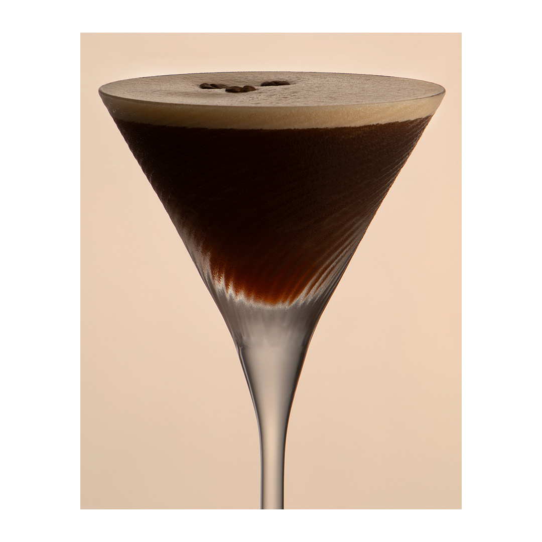 Close-up of an espresso martini in a clear glass with three coffee beans floating on top, set against a creamy beige background.