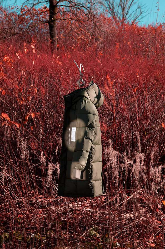 Padded olive green hooded jacket hanging on a metal hanger in an outdoor setting surrounded by tall red vegetation.