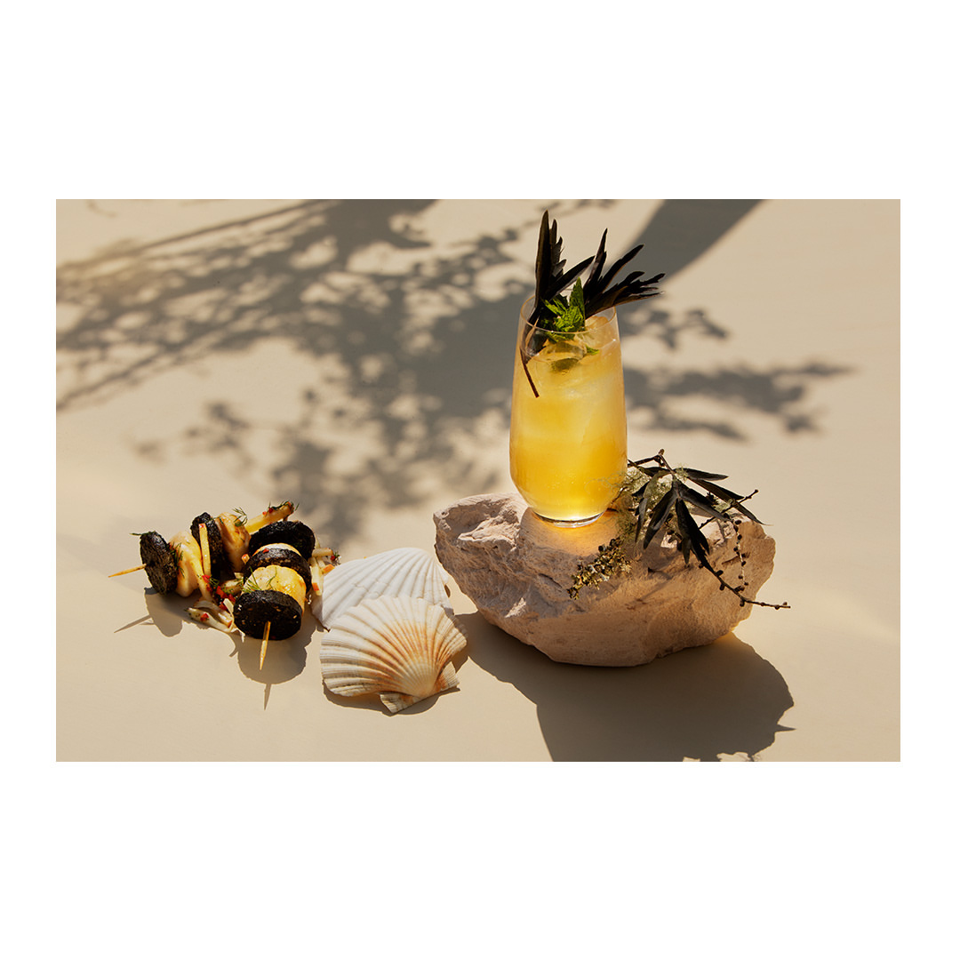 Refreshing yellow cocktail garnished with mint and dark sprigs on a light surface with seashells and a food skewer, lit by soft sunlight.