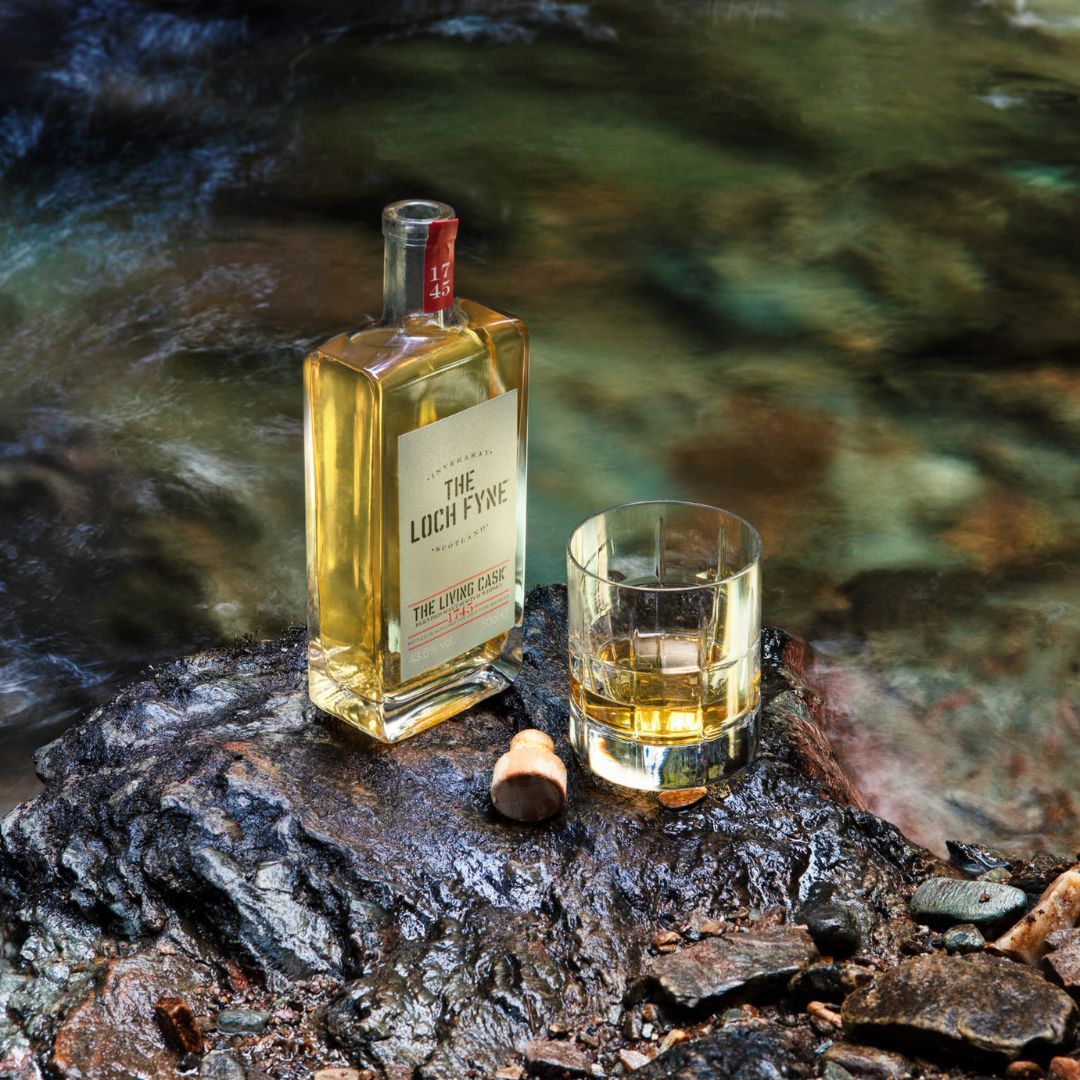 Whiskey bottle and glass on a rock near flowing water, showcasing the product in a natural setting with blurred motion.