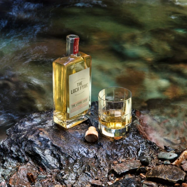 Whiskey bottle and glass on a rock near flowing water, showcasing the product in a natural setting with blurred motion.