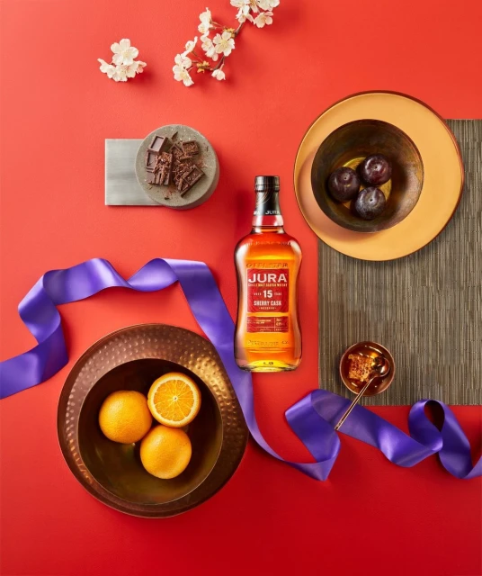 Still life featuring Jura whisky bottle, oranges, plums, chocolate, flowers, ribbon, and dishes against a red background.