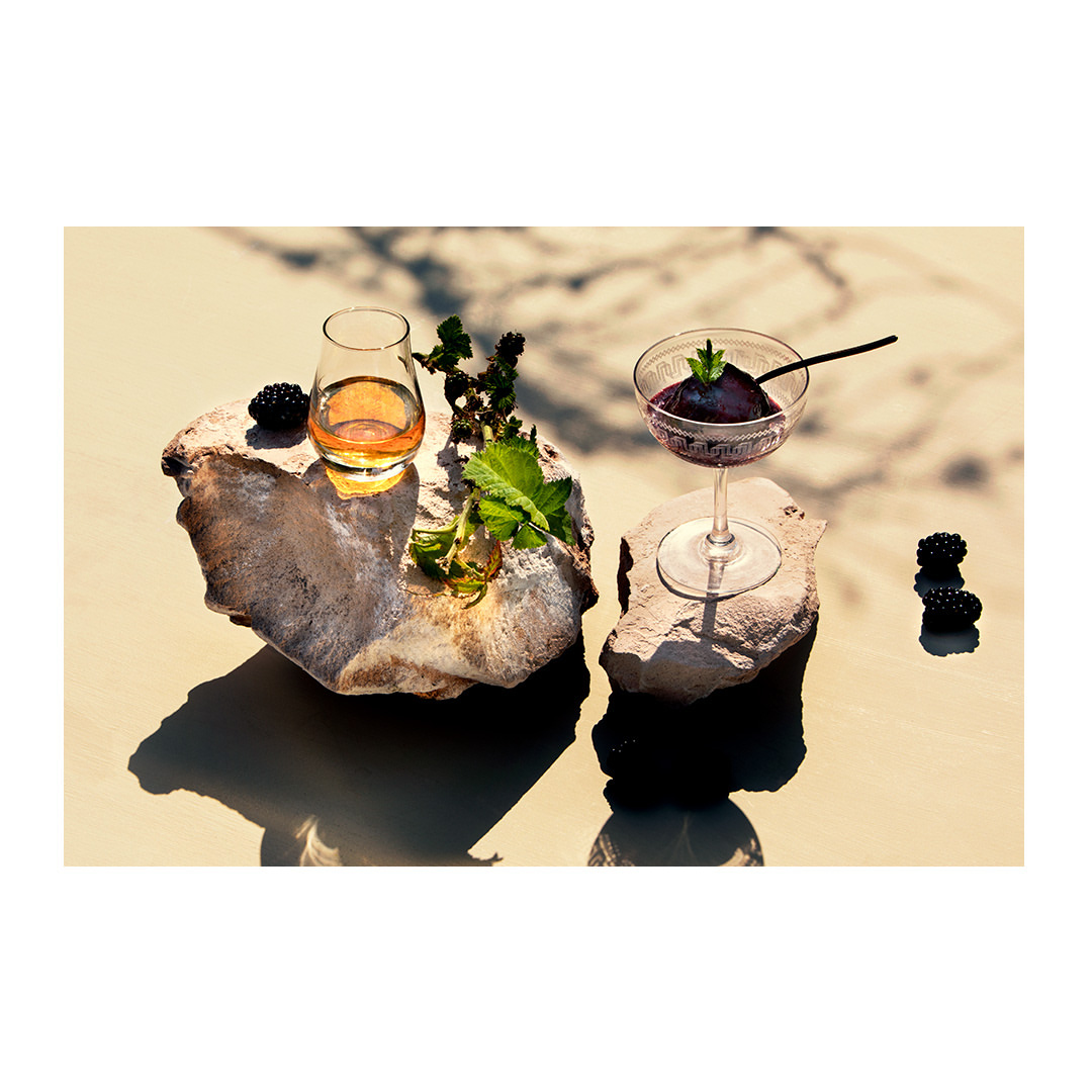 Two cocktails, one amber and the other purple, sit atop rocks with mint leaves and blackberries on a tan surface with shadows.