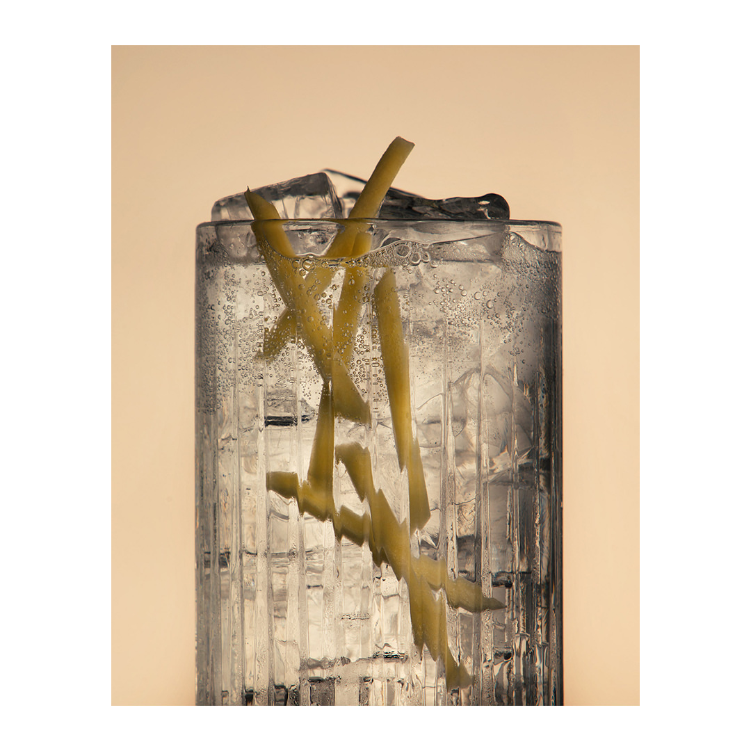 Tall ribbed glass filled with ice and sparkling tonic water, garnished with lemon zest, against a soft beige backdrop.