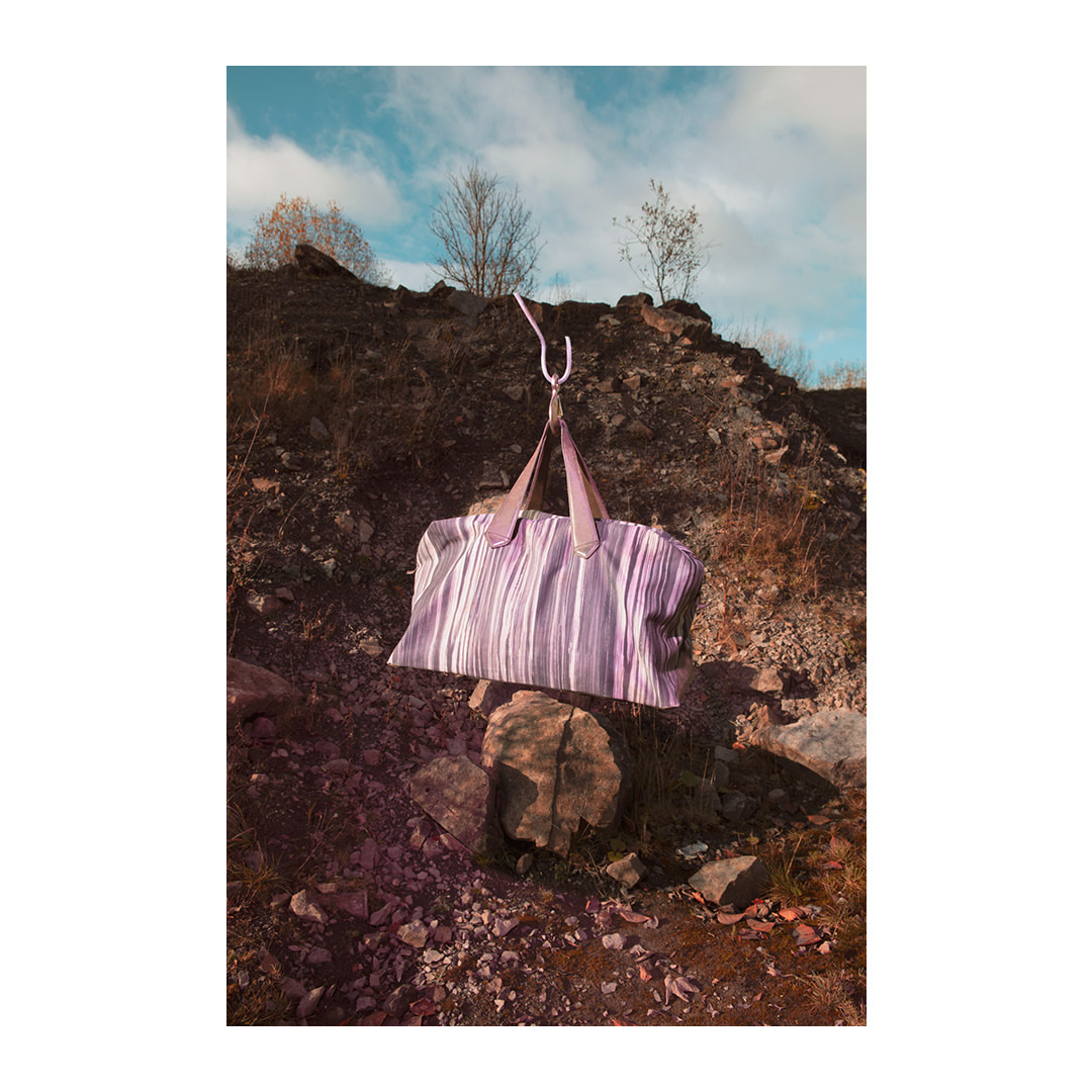 Tan-striped duffel bag hanging from a silver hook against a rocky hillside backdrop with sparse trees and a partly cloudy sky.