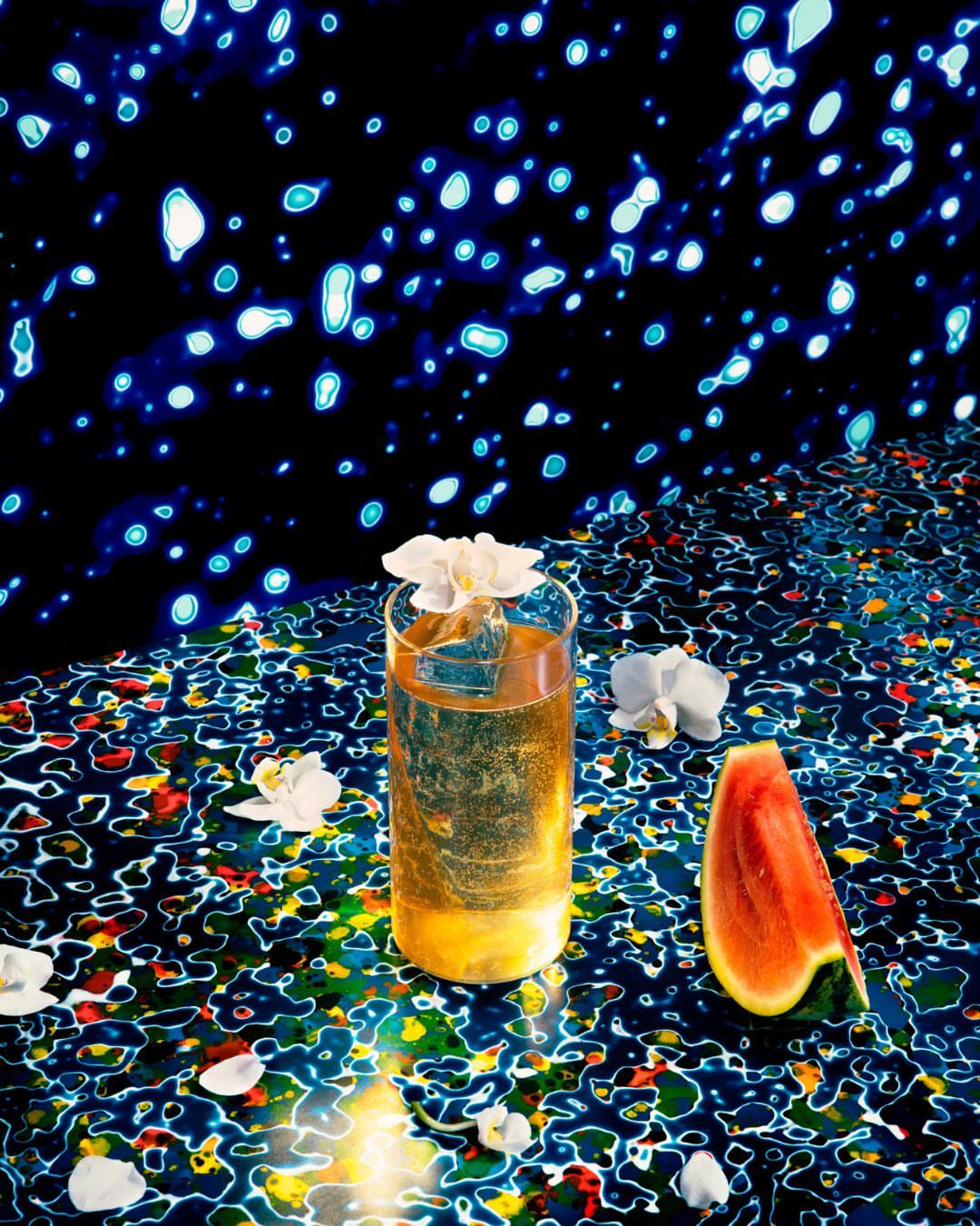 Studio shot of a cocktail in a glass with ice and a white flower garnish, plus watermelon, set against a vibrant, patterned backdrop.