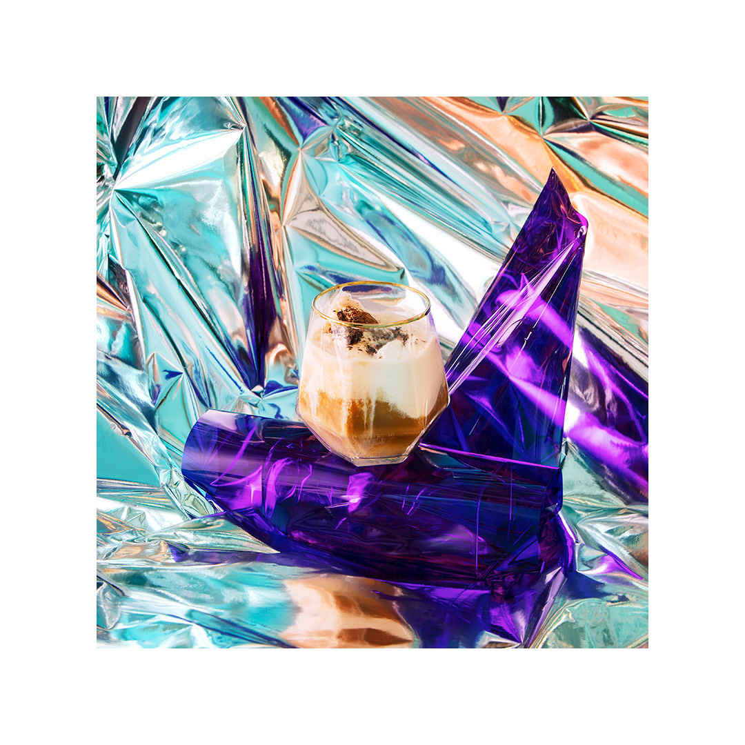 Dessert drink in clear glass sits beside folded purple material, turquoise and orange metallic backgrounds create a studio setting.