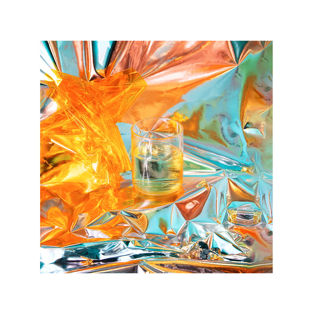 Crystal glass filled with liquid reflects vibrant copper and turquoise colors, set against a shimmering, crumpled metallic background.