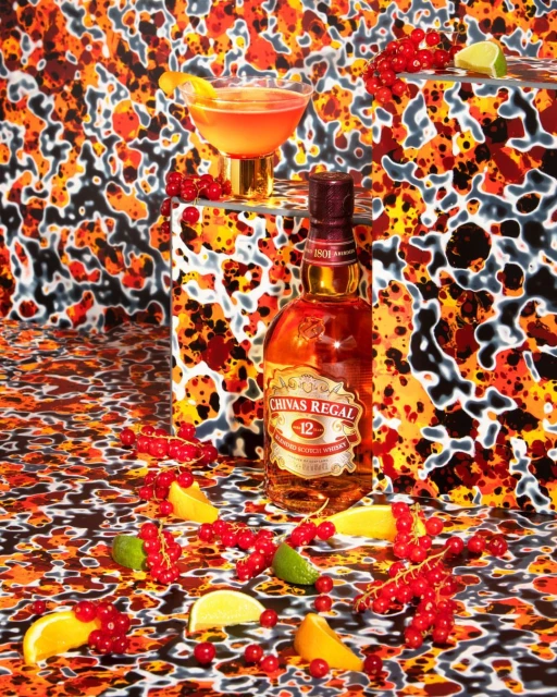 Still life shot of a bottle of Chivas Regal whisky, a cocktail glass, sliced citrus fruit, and red currants on a patterned surface.