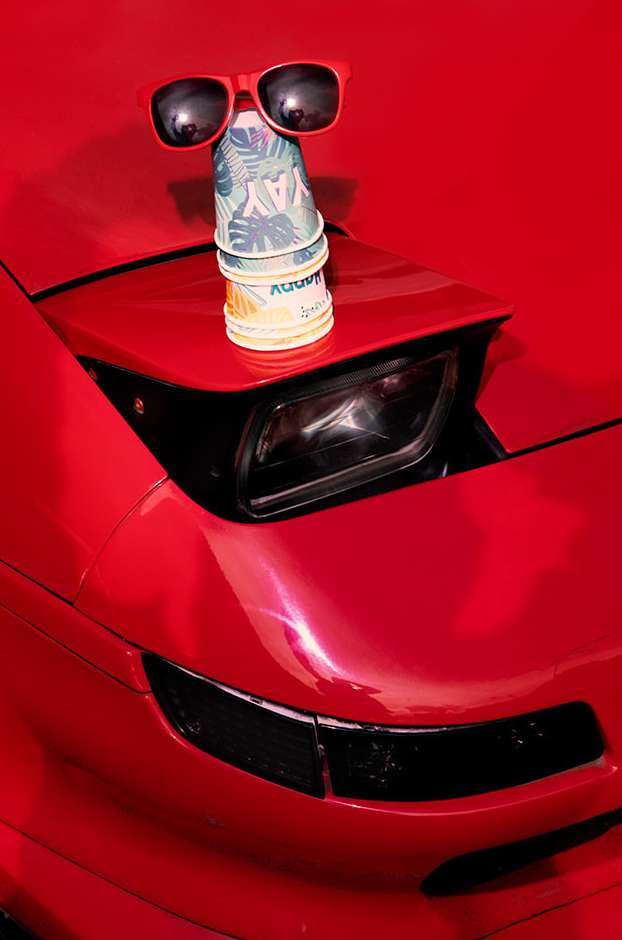 Close-up of a red car with sunglasses perched on top of stacked disposable cups, creating a whimsical face on the vehicle's hood.