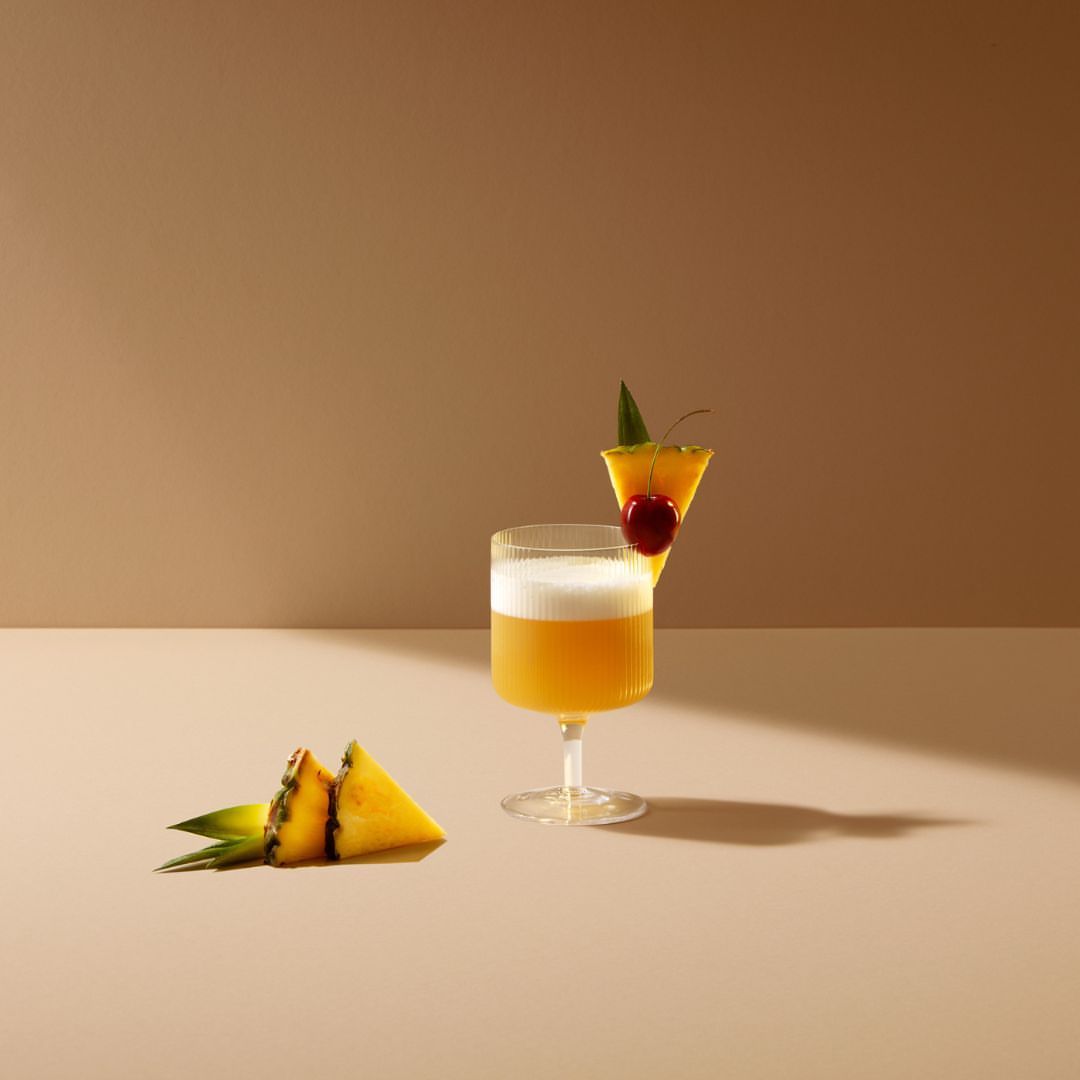 A cocktail in a stemmed glass is garnished with a pineapple slice and a cherry. Pineapple wedges lay near the base of the glass.