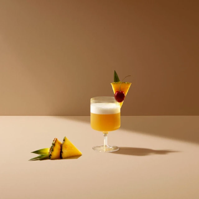 A cocktail in a stemmed glass is garnished with a pineapple slice and a cherry. Pineapple wedges lay near the base of the glass.