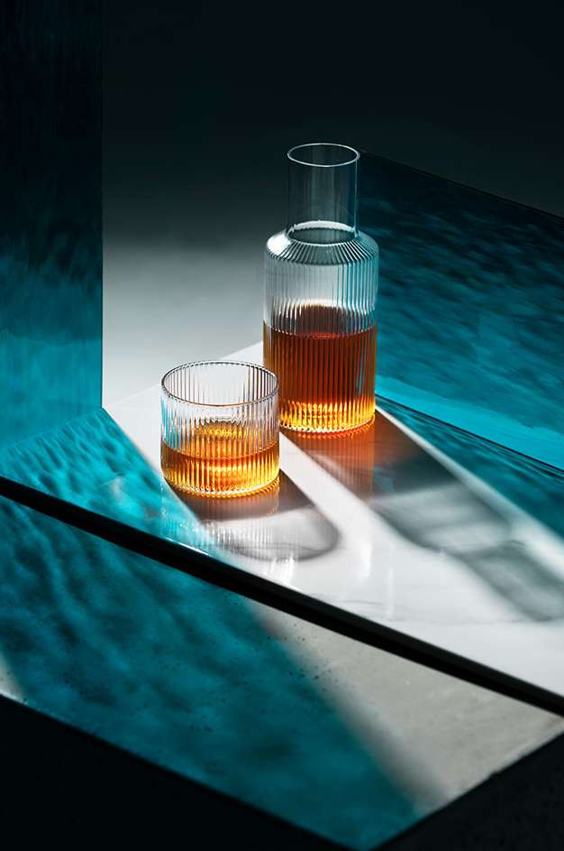 Crystal glass decanter and short glass, both filled with amber liquid, illuminated by a sharp white light against a blue background.