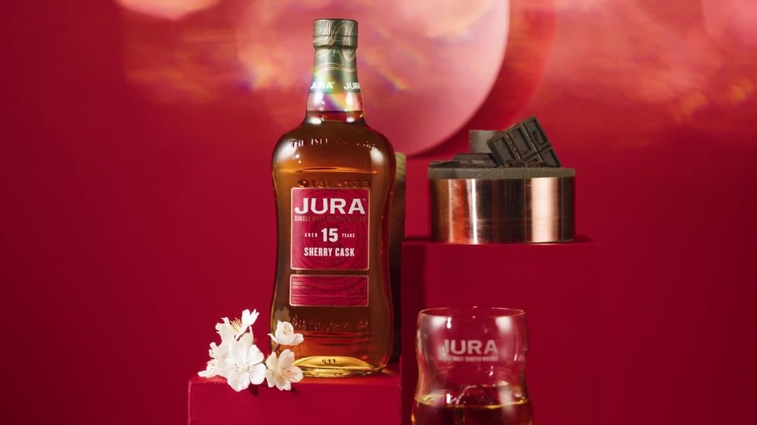 Jura 15 Year Sherry Cask single malt scotch bottle on a red platform with a glass and copper-trimmed slate pedestal.