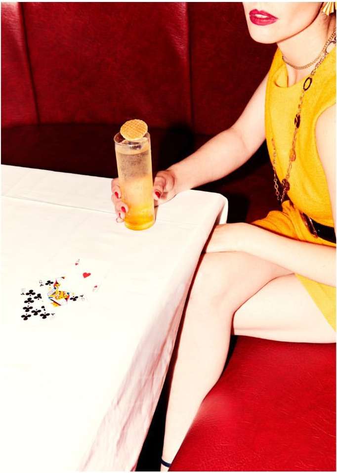 Stylish woman in yellow dress with cocktail, cards on table, and red booth seating. Red lipstick, gold jewelry, indoor setting.