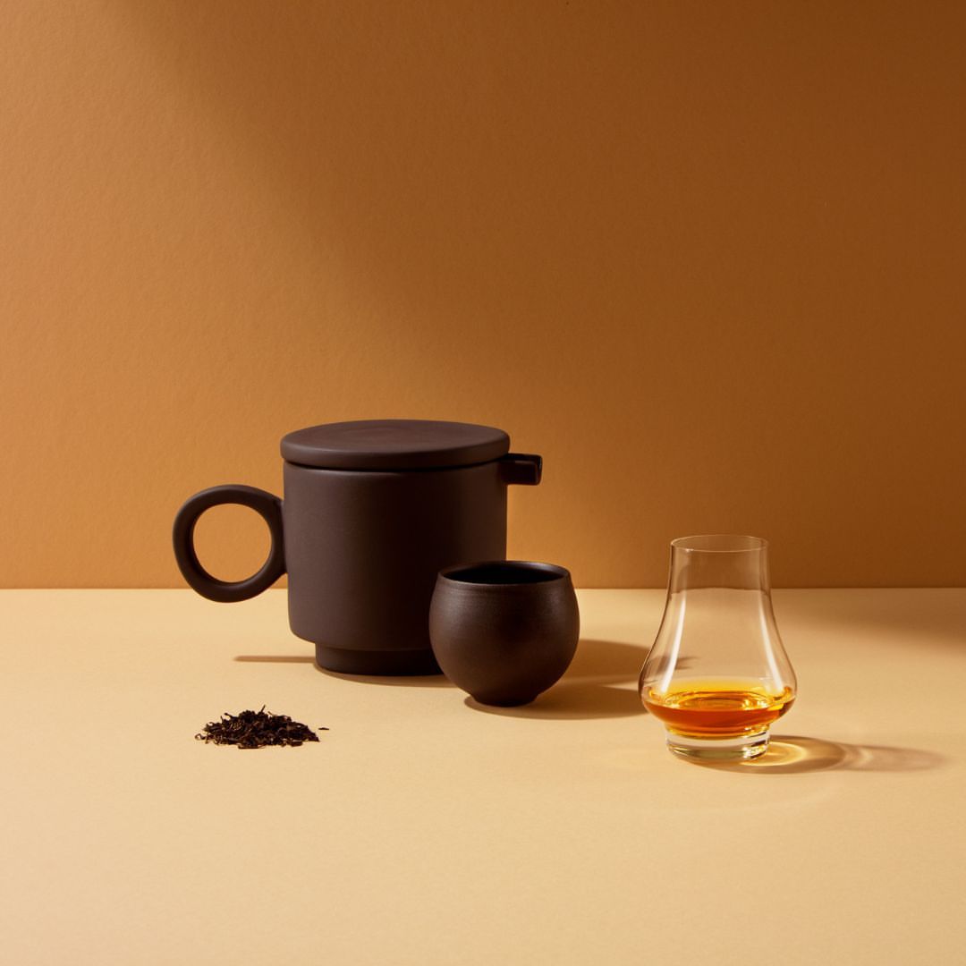 Studio shot of a dark brown tea set, including a mug with a lid, a small cup, and loose tea leaves, next to a Glencairn glass.