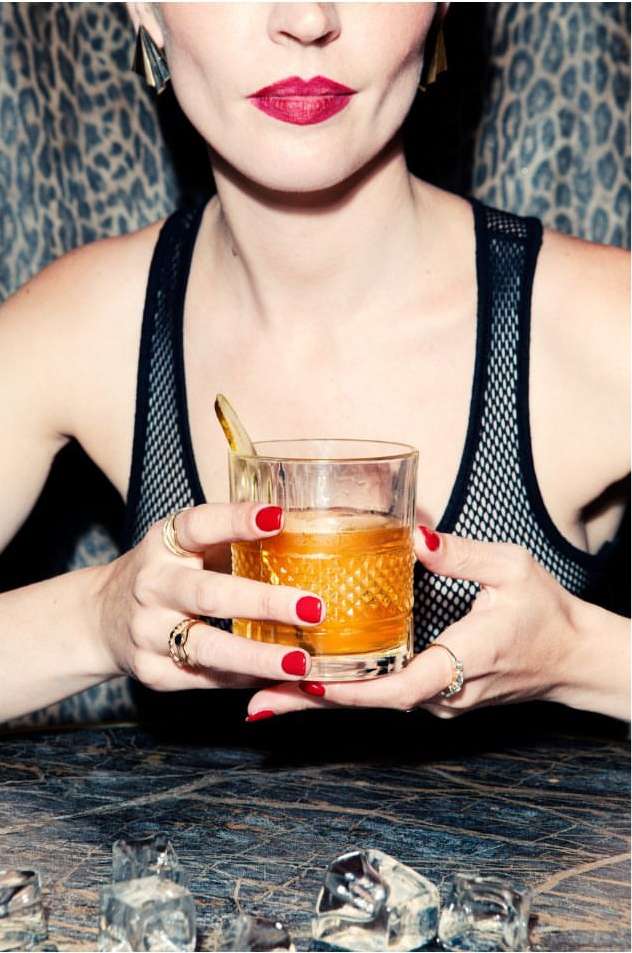 Adult woman holding cocktail glass with amber liquid, gold rings, and red nails, set against an animal print backdrop.