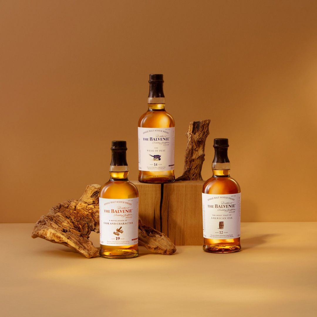 Three bottles of The Balvenie single malt Scotch whisky are arranged on wooden blocks and a piece of driftwood against a brown backdrop.
