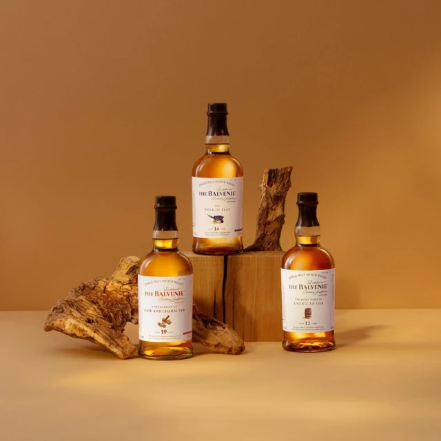 Three bottles of The Balvenie single malt Scotch whisky are arranged on wooden blocks and a piece of driftwood against a brown backdrop.