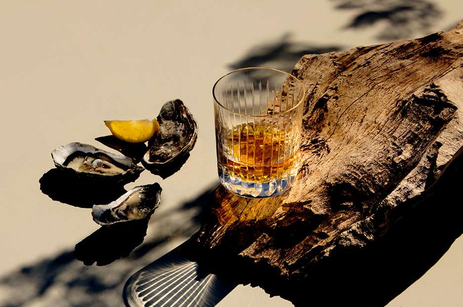 A glass of amber liquid, oysters, and a lemon wedge sit on a textured wood surface, evoking a sense of luxury and culinary delight.