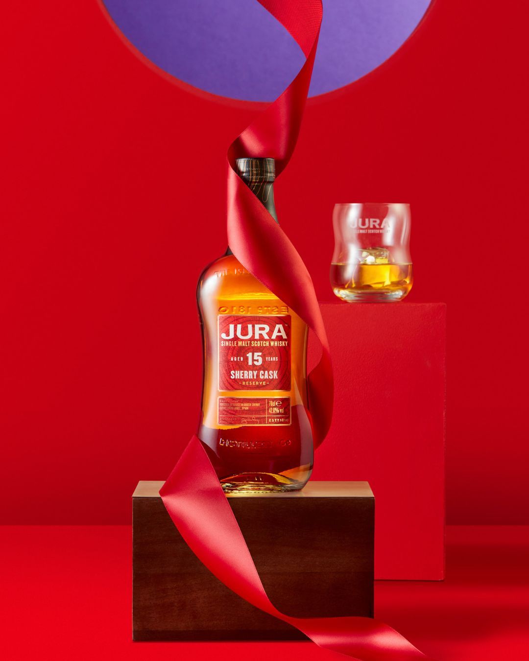Jura single malt Scotch whisky bottle with red ribbon displayed on wooden box against a red backdrop with glass and liquid.