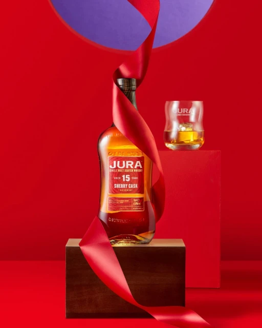 Jura single malt Scotch whisky bottle with red ribbon displayed on wooden box against a red backdrop with glass and liquid.