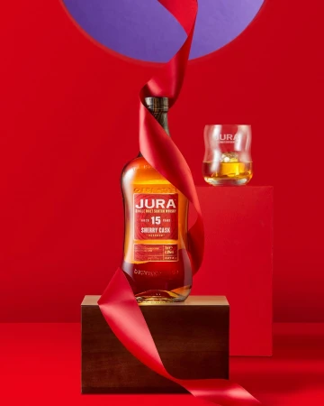 Jura single malt Scotch whisky bottle with red ribbon displayed on wooden box against a red backdrop with glass and liquid.