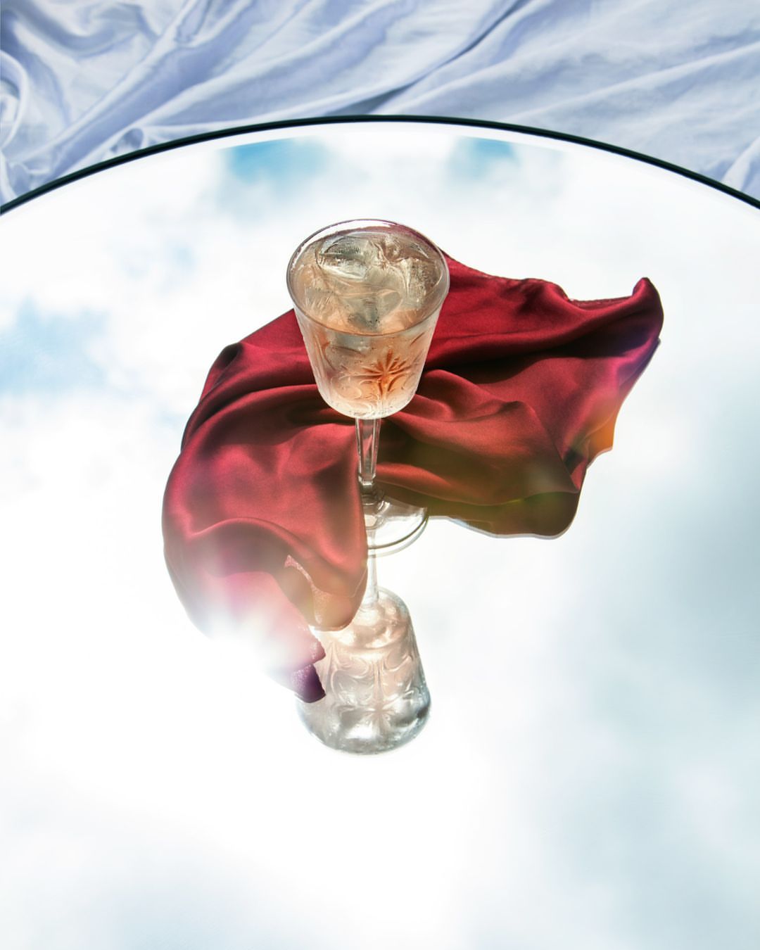Stemmed crystal glass with ice and a red cloth sits on a round mirror, reflecting the sky, against a white draped fabric background.