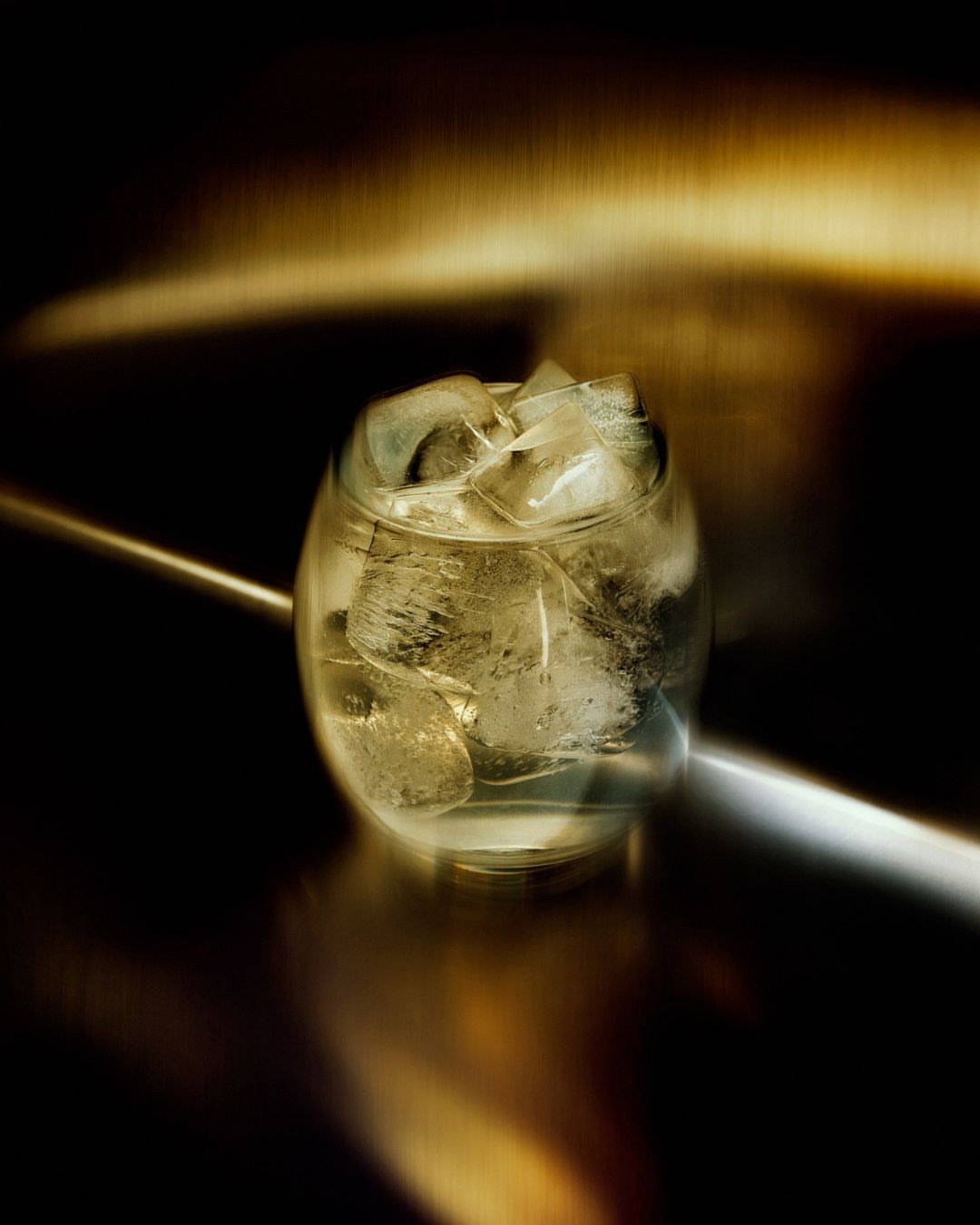 Close-up of a clear glass filled with ice cubes, illuminated by a golden light source, against a dark and reflective background.