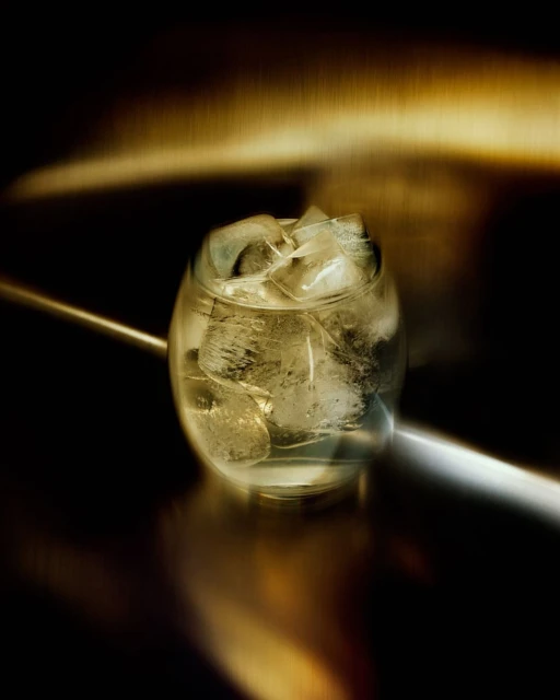 Close-up of a clear glass filled with ice cubes, illuminated by a golden light source, against a dark and reflective background.
