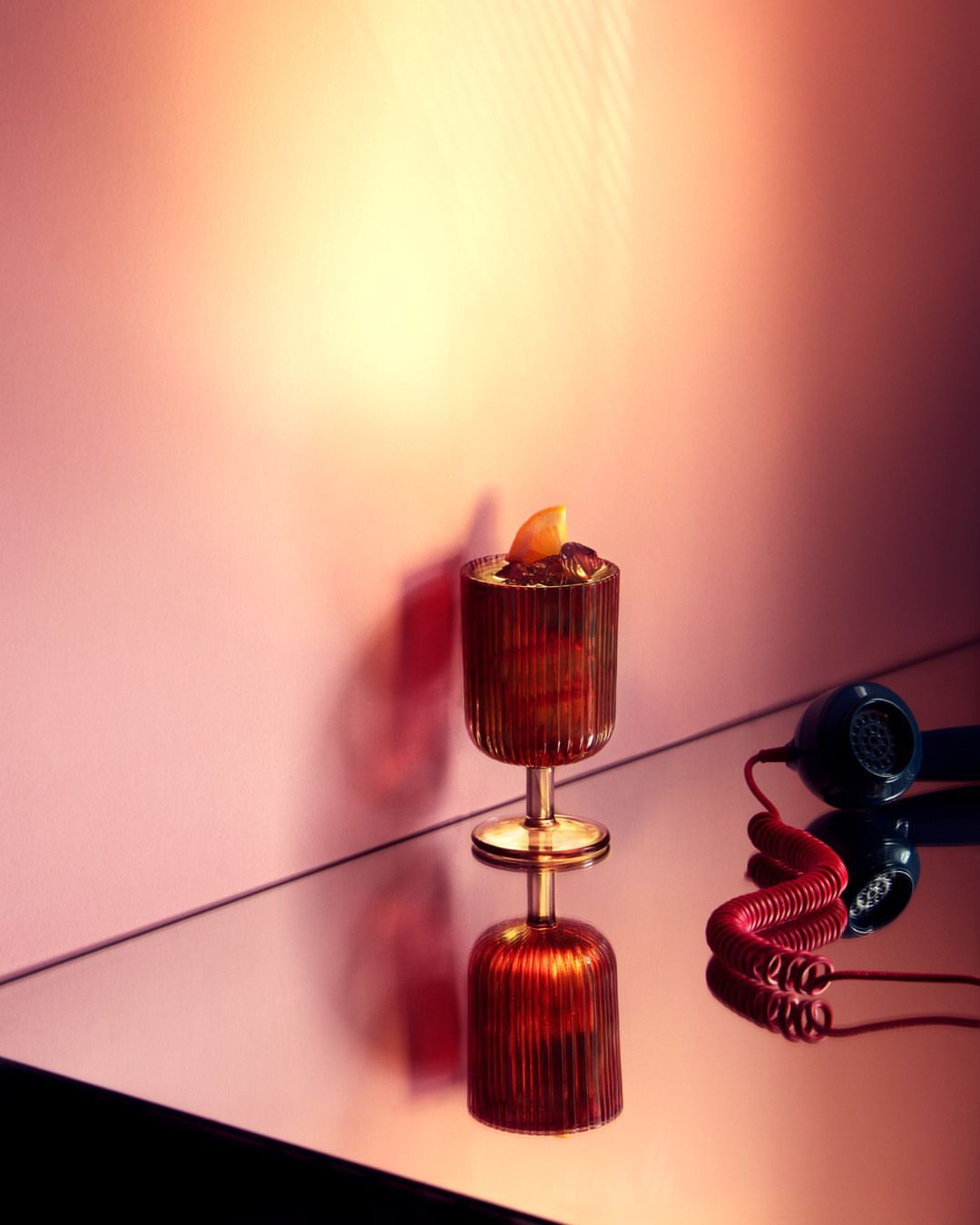 A cocktail in a gold-stemmed glass sits on a reflective surface, next to a set of headphones, against a pink-toned wall.