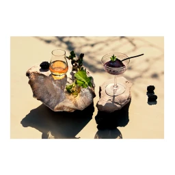Two cocktail glasses with amber liquid and dark berries atop stone slabs on a sandy surface in bright sunlight by Armando Ferrari.