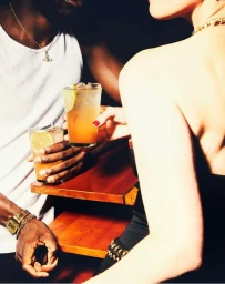 Close-up portrait of two people holding cocktail glasses with lime slices at a wooden bar, photographed by Armando Ferrari.