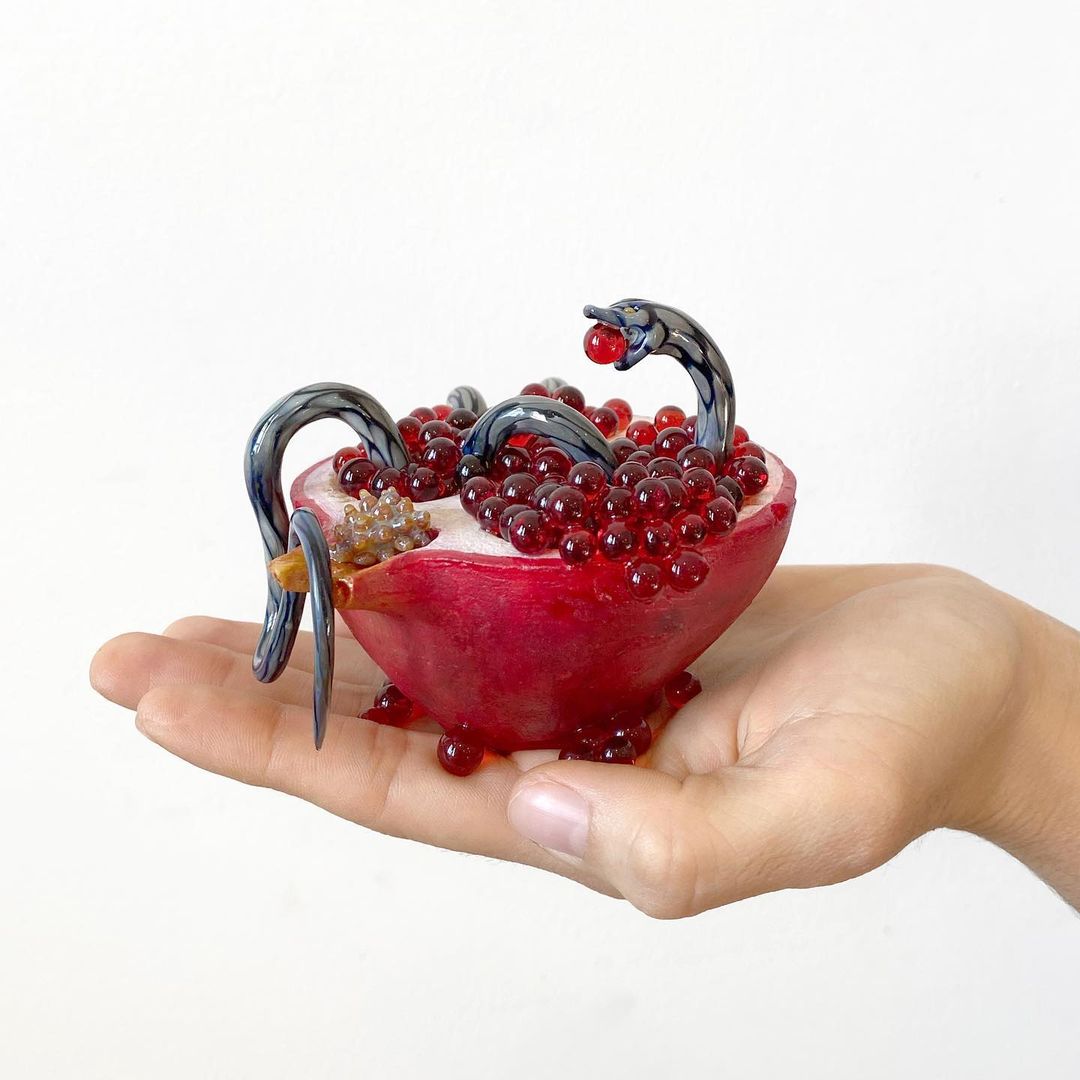 Hand holding a halved pomegranate with glass snake sculptures feasting on the red arils, set against a bright white background.