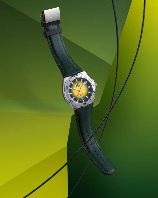 Wristwatch with a silver casing and a yellow face with a green leather band floating on a green geometric background in a studio.
