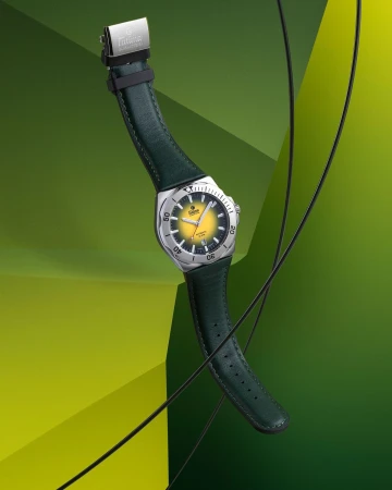 Wristwatch with a silver casing and a yellow face with a green leather band floating on a green geometric background in a studio.