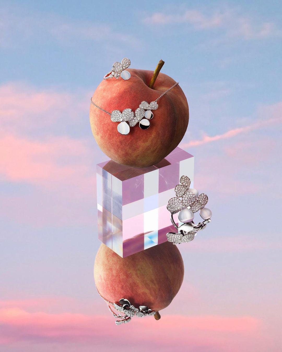 Elevated view of a silver necklace and ring with a floral pattern on a peach, balanced on a translucent block against a blue and pink sky.
