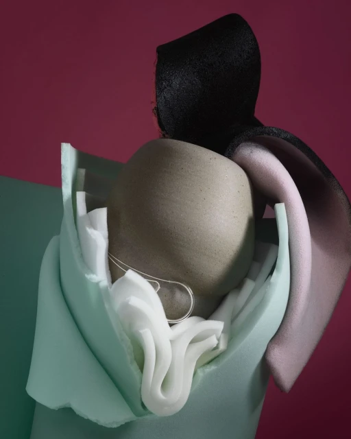Abstract still life with a tan human head-shaped form, a black hat, and soft mint and white fabric textures against a red backdrop.