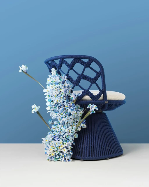 Woven blue accent chair with a light cushion and a floral accent piece against a graduated blue background in a studio setting.