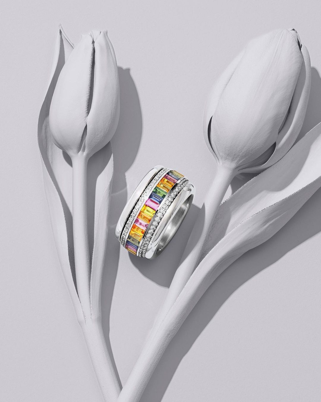 Close-up of a silver ring with multicolored gemstones next to two closed, white tulip blossoms on a light gray surface.