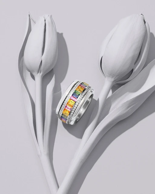 Close-up of a silver ring with multicolored gemstones next to two closed, white tulip blossoms on a light gray surface.